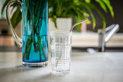 CaliWoods Reusable Drinking Straw made from stainless steel and with a bend. Straw is in a glass and there's a blue vase in the background. 